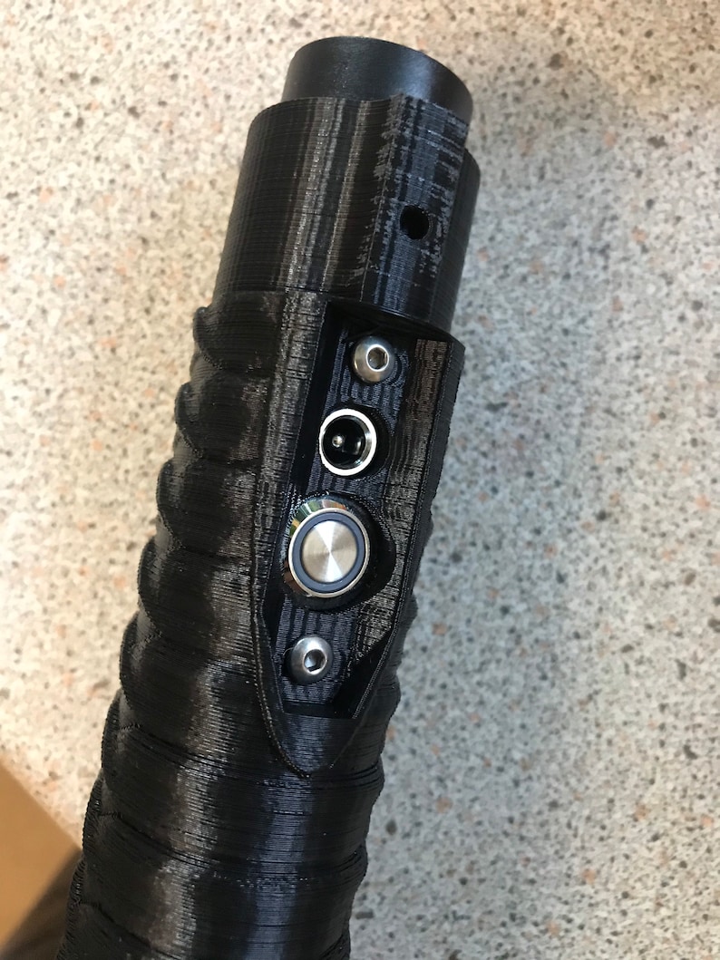 Puede incluir: Primer plano de una empu&ntilde;adura de sable l&aacute;ser negra con un bot&oacute;n plateado y un conector de cable de alimentaci&oacute;n negro.