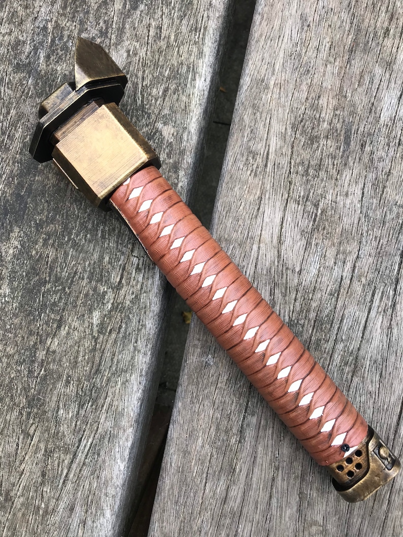 Puede incluir: Una espada samur&aacute;i marr&oacute;n y blanca con una empu&ntilde;adura de metal dorado. La empu&ntilde;adura tiene un dise&ntilde;o &uacute;nico con una base cuadrada y una parte superior puntiaguda.