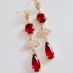 May include: Gold-toned dangle earrings with red oval and teardrop-shaped gemstones. The earrings feature fan-shaped accents with small clear stones. The earrings have a decorative filigree design at the top.