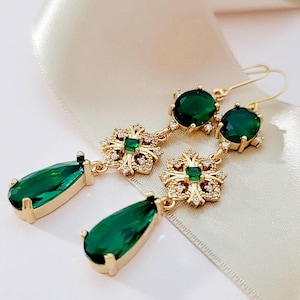 May include: A pair of gold-tone drop earrings featuring a round green gemstone at the top, a central square-cut green stone surrounded by small clear crystals in a decorative frame, and a large pear-shaped green gemstone at the bottom.