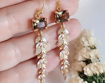 May include: Gold-toned dangle earrings featuring rectangular brown gemstones at the top. The earrings have a leaf-like design with clear, sparkling accents. The earrings are designed with a hook closure.