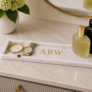 May include: A rectangular white marble tray featuring the gold-colored letters ARW printed in the center. The tray holds a gold-toned bangle, a silver and gold wristwatch with a clover charm, a clear glass perfume bottle, and a black perfume bottle.
