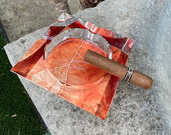 May include: A square glass ashtray with a red and orange leaf design. A brown cigar is resting on the edge of the ashtray.