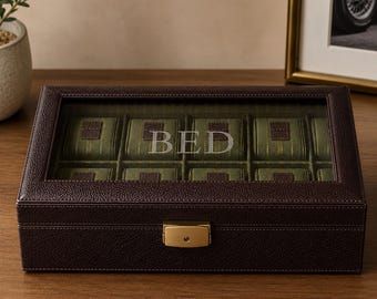 May include: A rectangular dark brown watch box with a textured exterior and a clear glass lid. The interior features green fabric-covered cushions for eight watches, with the letters BED etched in white on the glass lid and a gold metal latch.