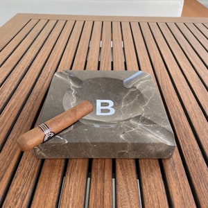 Pu&ograve; includere: Un posacenere in marmo marrone con una lettera "B" bianca al centro. Un sigaro marrone &egrave; appoggiato sul bordo del posacenere. Il posacenere &egrave; su un tavolo di legno.