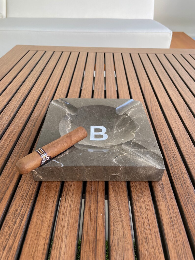 Pu&ograve; includere: Un posacenere in marmo marrone con una lettera "B" bianca al centro. Un sigaro marrone &egrave; appoggiato sul bordo del posacenere. Il posacenere &egrave; su un tavolo di legno.