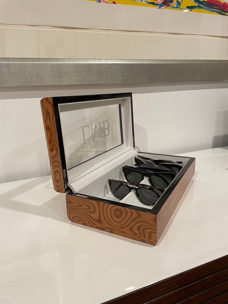 K&ouml;nnte beinhalten: Eine Holzkiste mit Glasdeckel und einer wei&szlig;en Aufschrift "GWB". Die Kiste ist ge&ouml;ffnet und enth&auml;lt vier Sonnenbrillen.