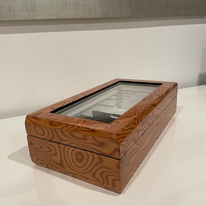 K&ouml;nnte beinhalten: Eine Holzuhrbox mit Glasdeckel. Die Box hat eine Holzmaserung und ist braun.