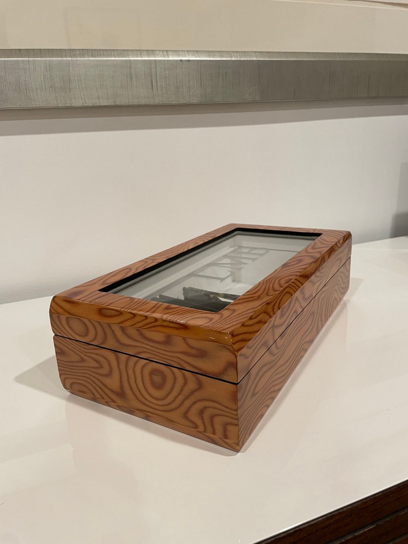 K&ouml;nnte beinhalten: Eine Holzuhrbox mit Glasdeckel. Die Box hat eine Holzmaserung und ist braun.