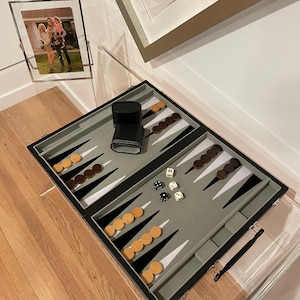 May include: A black and gray backgammon game set with a leather carrying case. The game board is open and shows the pieces in play. The dice are on the board and show the numbers 4 and 6.