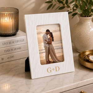 May include: A vertical rectangular photo frame with a white wood grain texture and a gold embossed G+D monogram at the bottom. The frame holds a photograph of a couple embracing on a beach at sunset, featuring a black easel back support.