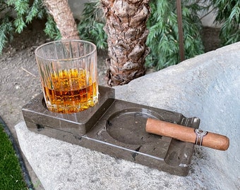 May include: A gray marble tray with two compartments, one for a glass of amber liquid and the other for a cigar. The tray is sitting on a light gray stone surface.