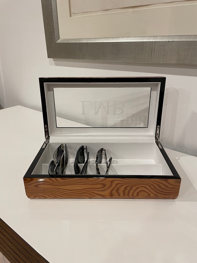 K&ouml;nnte beinhalten: Eine Holzkiste mit einem schwarzen Deckel und einer transparenten Glasplatte. Die Kiste hat eine Holzmaserung und der Deckel ist mit den Initialen "GWB" graviert. Die Kiste ist ge&ouml;ffnet und enth&auml;lt vier Sonnenbrillen.