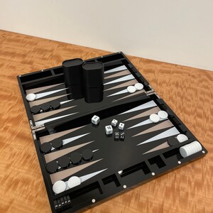 May include: A black backgammon set with white and tan playing pieces. The set is open and shows the playing board with dice and cups.