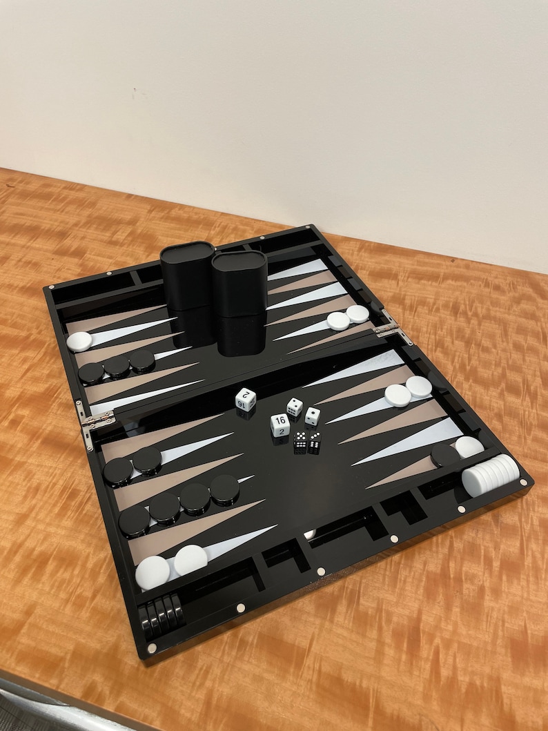May include: A black backgammon set with white and tan playing pieces. The set is open and shows the playing board with dice and cups.