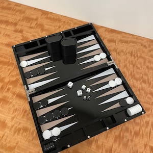 May include: A black backgammon set with white and tan triangles, black and white checkers, and dice. The set is open and ready to play.