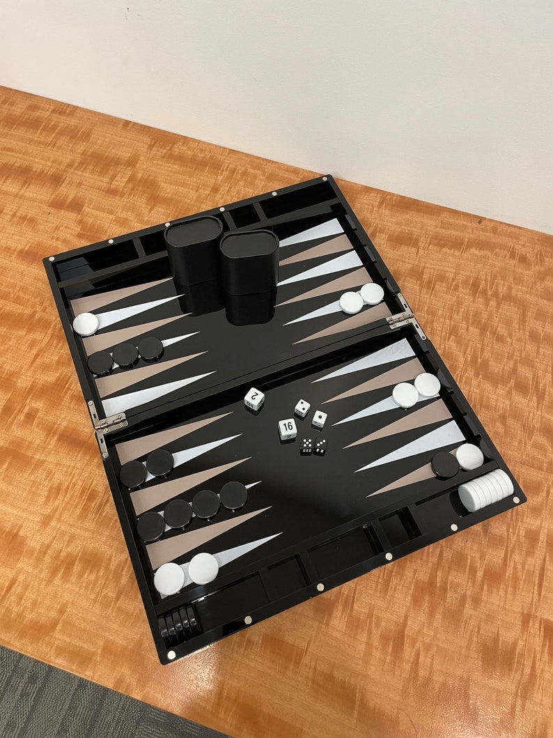 May include: A black backgammon set with white and tan triangles, black and white checkers, and dice. The set is open and ready to play.