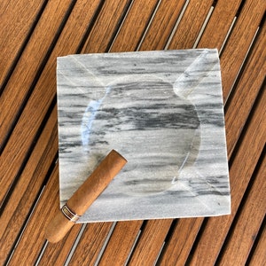 Pu&ograve; includere: Un portacenere in marmo grigio con un sigaro marrone appoggiato sopra. Il portacenere &egrave; su una superficie di legno.