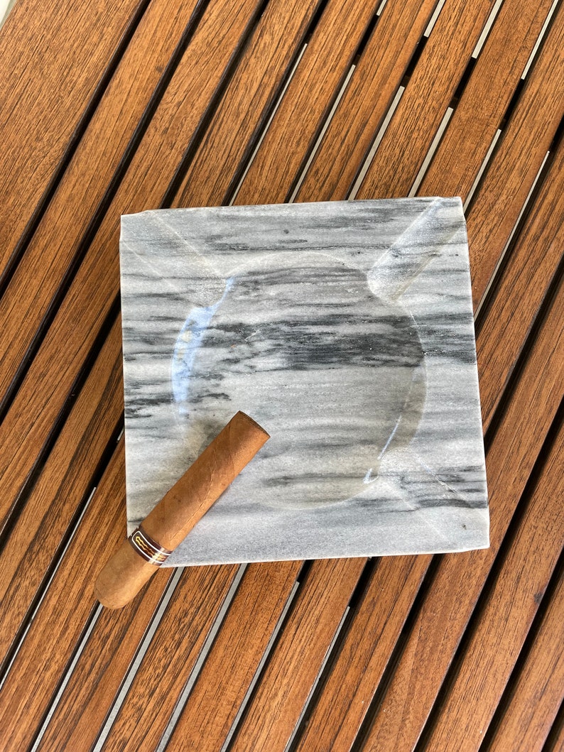 Pu&ograve; includere: Un portacenere in marmo grigio con un sigaro marrone appoggiato sopra. Il portacenere &egrave; su una superficie di legno.