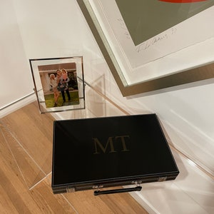 May include: A black leather game case with a gold embossed "MIT" logo and a silver handle. The case is sitting on a wooden floor.