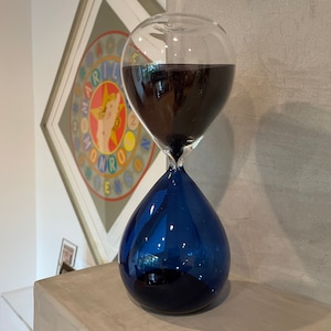 May include: A clear glass hourglass with dark liquid in the top bulb, connected to a deep blue, teardrop-shaped base. The hourglass is set on a concrete surface, with a framed artwork in the background.
