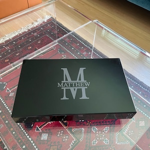 May include: A black rectangular box with a gray monogram of the letter "M" and the name "MATTHEW" engraved on the top.