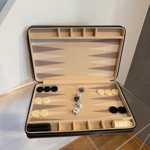 May include: An open backgammon board game set featuring a dark brown exterior case with a beige interior playing surface. The board includes light and dark brown triangular points, black and white circular checkers, and four dice, one marked with the number 2.