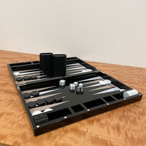 May include: A black backgammon set with white and black playing pieces, two dice, and two black containers. The set is open and ready to play.