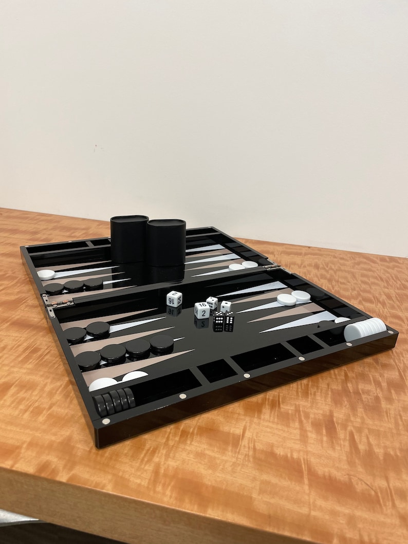 May include: A black backgammon set with white and black playing pieces, two dice, and two black containers. The set is open and ready to play.