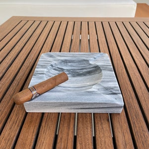 Pu&ograve; includere: Un posacenere in marmo grigio con un sigaro appoggiato sul bordo. Il posacenere &egrave; su un tavolo di legno.