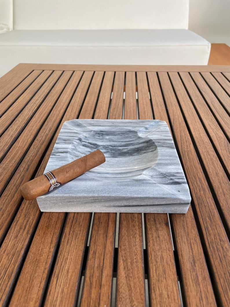 Pu&ograve; includere: Un posacenere in marmo grigio con un sigaro appoggiato sul bordo. Il posacenere &egrave; su un tavolo di legno.