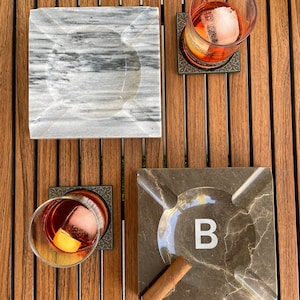 Pu&ograve; includere: Due posacenere quadrati in marmo, uno grigio e uno marrone con la lettera "B" al centro. Un sigaro marrone e due bicchieri con cubetti di ghiaccio e un liquido rosso sono su una superficie di legno.