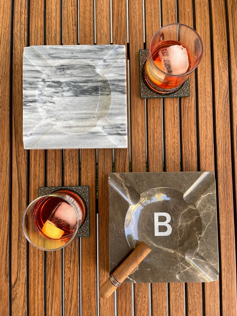 Pu&ograve; includere: Due posacenere quadrati in marmo, uno grigio e uno marrone con la lettera "B" al centro. Un sigaro marrone e due bicchieri con cubetti di ghiaccio e un liquido rosso sono su una superficie di legno.