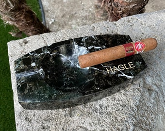 May include: A black marble ashtray with a gold inscription that reads "HAGLE". A brown cigar is resting on the edge of the ashtray.