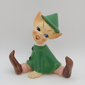 May include: A ceramic figurine of a green elf with a pointed hat, sitting with legs crossed and wearing brown boots. The elf has a light brown face with blue eyes and a red mouth.