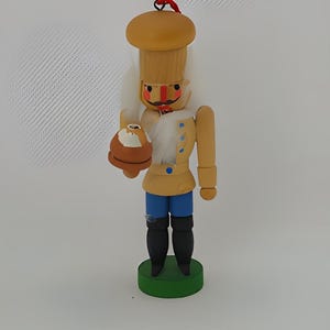 May include: A wooden nutcracker figurine with a brown hat, white beard, blue pants, and a red and white striped collar. The nutcracker is holding a brown nut in its left hand.