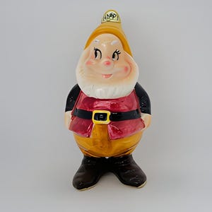 May include: A ceramic figurine of a dwarf from the Disney movie Snow White and the Seven Dwarfs. The dwarf is wearing a yellow hat with the word "Happy" on it, a red shirt with a black belt, and yellow pants. He has a friendly smile and is standing with his hands in his pockets.