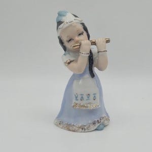 May include: A ceramic figurine of a girl playing a flute. The figurine is dressed in a light blue dress with a white apron and a matching hat. The flute is gold-colored, and the figurine has black hair.