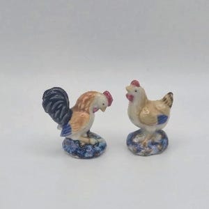 May include: Two ceramic figurines of a rooster and a hen. The rooster has a black and blue tail, brown and white body, and red comb. The hen is beige with blue accents and a red comb. Both are perched on blue and white bases.