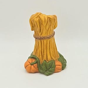 May include: A decorative figurine of a wheat sheaf, with a brown rope around the middle. The base features two orange pumpkins and green leaves. The figurine is painted in shades of yellow, orange, and green.