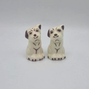 May include: Two ceramic dog figurines. The dogs are white with brown ears and black spots on their paws. They have black facial features and a Y-shaped marking on their chests. The figurines are sitting upright.