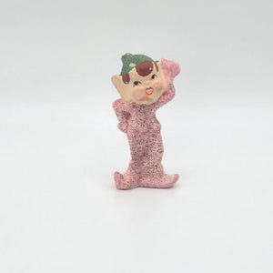 May include: A vintage figurine of a whimsical elf with a pink glittery body and a green and brown hat. The elf has a smiling face with rosy cheeks and is posed with one hand on its head.