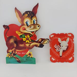 May include: Two vintage Valentine's Day cards featuring cartoon donkeys. The first card is a pop-up card with a brown donkey holding a red heart that says "Be Mine". The second card is a flat card with a white donkey wearing a red ribbon. Both cards have a red heart shape and are decorated with white hearts.