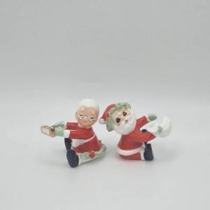 May include: Two ceramic Christmas figurines. One is a seated Mrs. Claus in a red and white dress, holding a book. The other is a seated Santa Claus in a red and white suit, holding a sack. Both have black shoes.