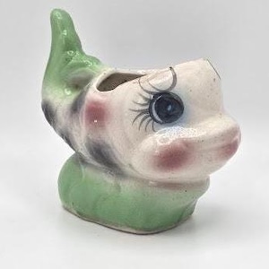 May include: A vintage ceramic planter shaped like a whimsical elf face. The planter features a green pointed hat, rosy cheeks, and large blue eyes with long eyelashes. The base and hat are green, with the face in off-white.