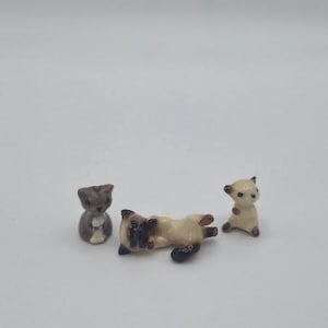 May include: Three miniature ceramic animal figurines. One is a small brown creature sitting upright, one is a cream-colored cat lying on its back with dark brown markings, and one is a small cream-colored creature standing on its hind legs.
