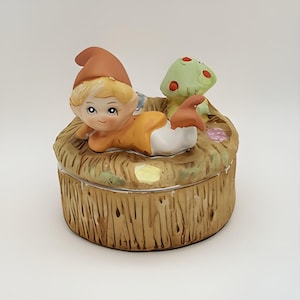 May include: A ceramic trinket box featuring a small elf figurine lying on a bed of straw with a mushroom beside it. The box is decorated with flowers and has a brown wood-like finish.