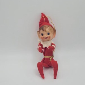 May include: A vintage Christmas elf ornament. The elf is dressed in a red outfit with white trim, a matching red hat, and a white collar. The elf has a smiling face with blue eyes and a gold string for hanging.