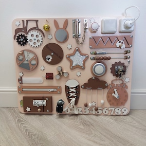 Könnte beinhalten: Ein rosa-braunes Holz-Beschäftigungsbrett mit verschiedenen Aktivitäten für Kinder. Es verfügt über einen Traktor, einen Spiegel, einen Stern, einen Spinner, einen Reißverschluss, ein Schloss, einen Lichtschalter, eine Uhr, eine Wolke mit hängenden Perlen, einen Schuh mit Schnürsenkeln, eine Kette, einen Schieber, eine Zahlenreihe und eine Glocke.