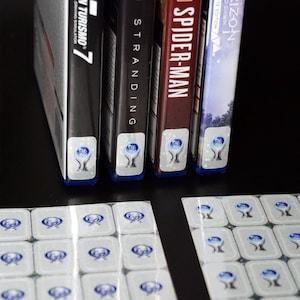 May include: Four video game cases with holographic stickers on the bottom. The cases are for the games Horizon Zero Dawn, Marvel's Spider-Man, Death Stranding, and Gran Turismo 7. The cases are stacked on top of each other. Two sheets of holographic stickers are on the table below the cases.
